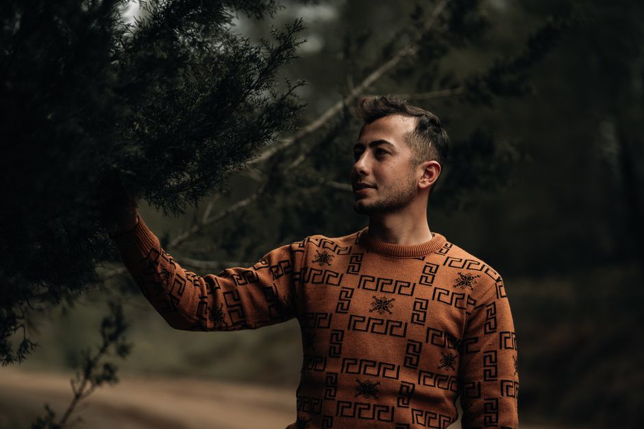 A man in a stylish sweater poses with a tree branch in a natural outdoor setting.