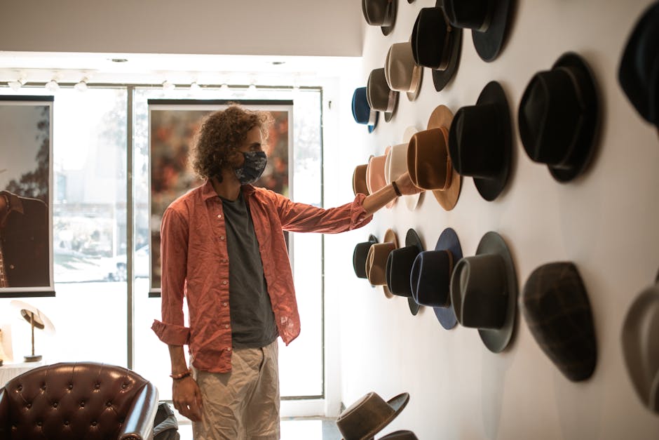 A man wearing a mask shops for hats in a boutique, touching various styles on display.