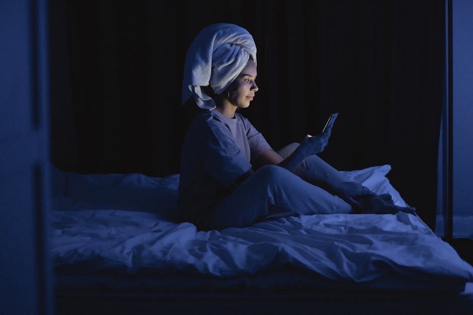 A woman sits on her bed with a towel on her head, engrossed in her smartphone in a dark room.