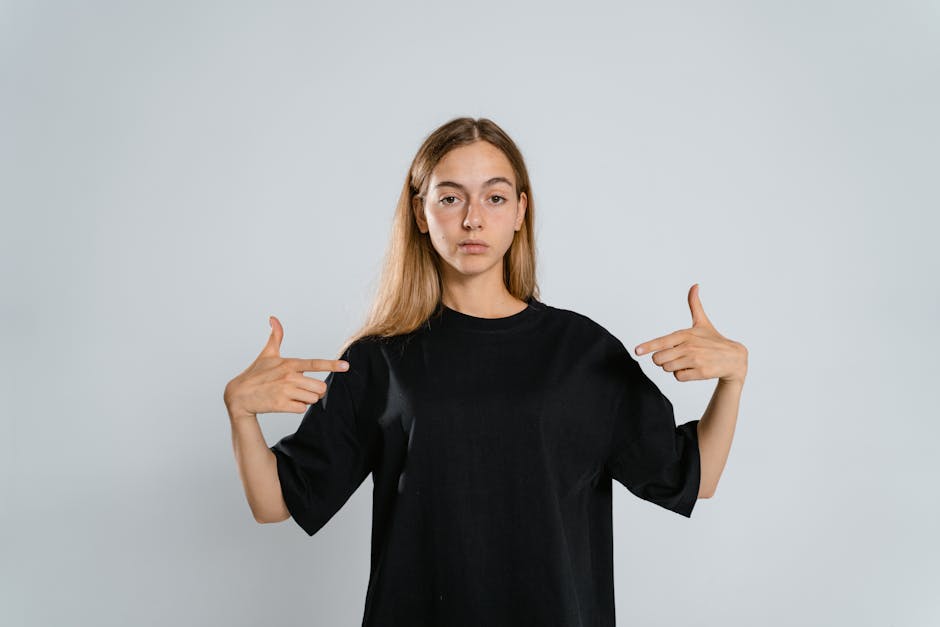 Portrait of a woman in a black t-shirt pointing at herself on a white background.