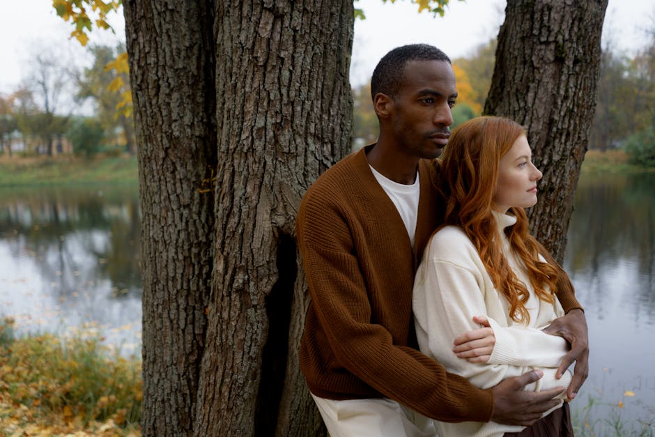 A diverse couple embracing by a lake with autumn leaves and trees.