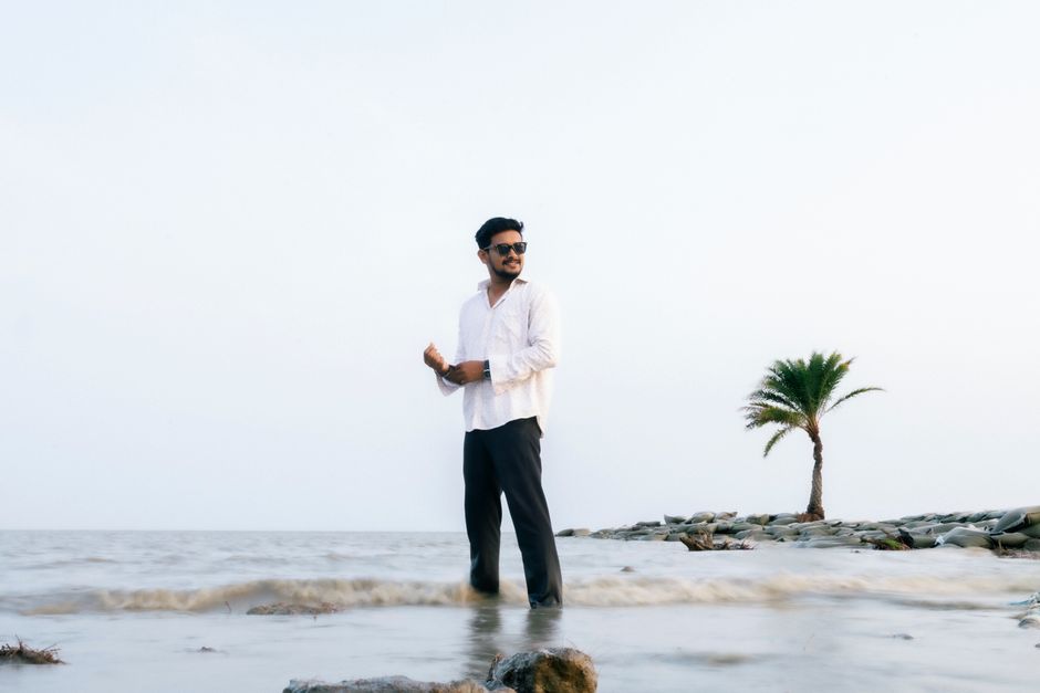 Man standing confidently on a rocky beach surrounded by waves and a lone palm tree, embracing nature.