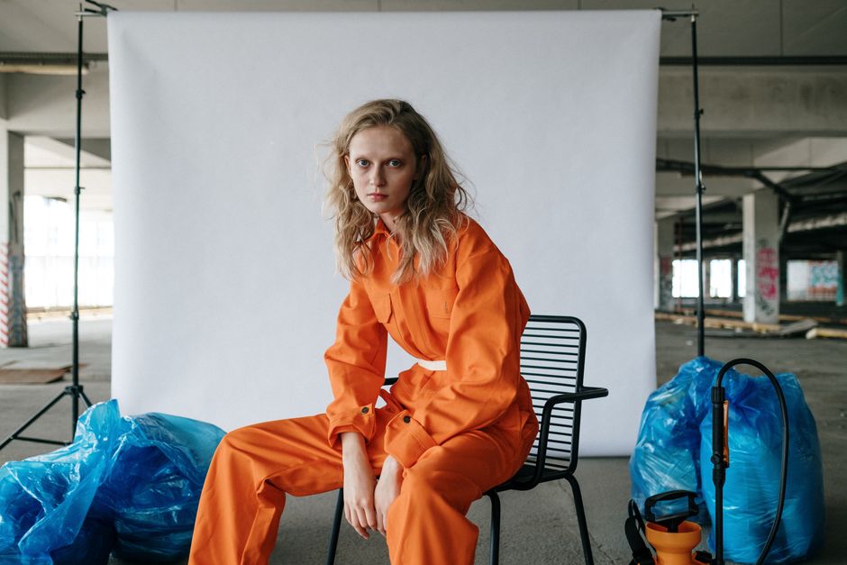 Stylish woman in orange suit seated indoors, surrounded by blue trash bags, exuding a modern, edgy vibe.