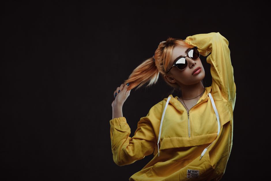 Fashionable woman wearing sunglasses and yellow hoodie posing confidently on a black background.