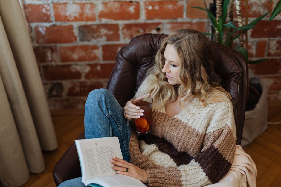 A woman enjoying a book and a cup of tea in a stylish armchair against a brick wall.