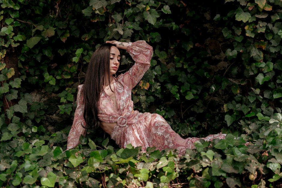 A fashionable woman in a floral dress poses elegantly among lush green ivy outdoors.