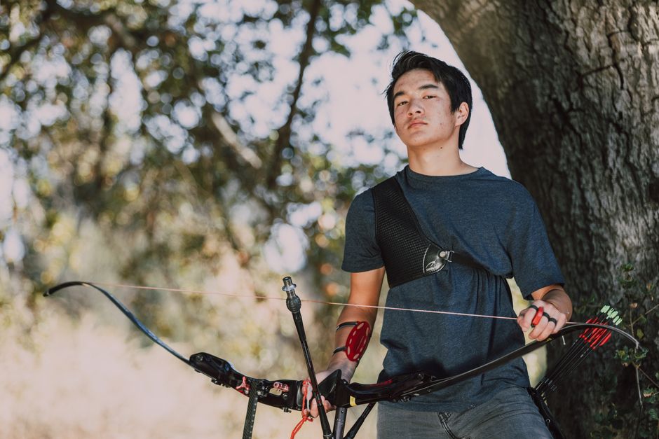 Teen male archer outdoors with bow and arrows in a sunny park setting.