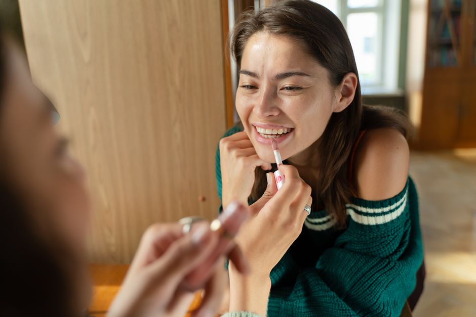 Young woman applying lipstick while smiling at her reflection inside a cozy room.